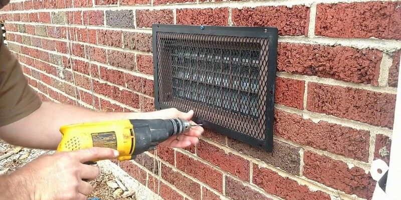 A man using a yellow drill to install a black galvanized steel eight-inch by sixteen-inch foundation vent cover on the side of a brick wall.