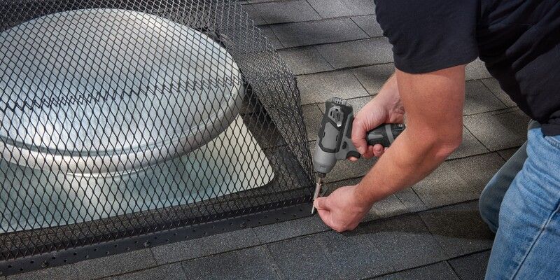 A man using a gray drill to install a black galvanized steel roof vent guard on a roof over a turtle-style roof vent.
