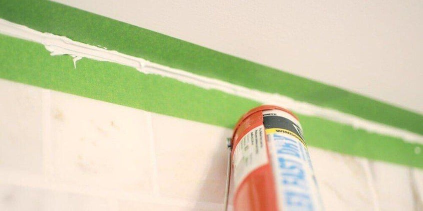 A caulk gun being used to fill in grout between two strips of green painter's tape.