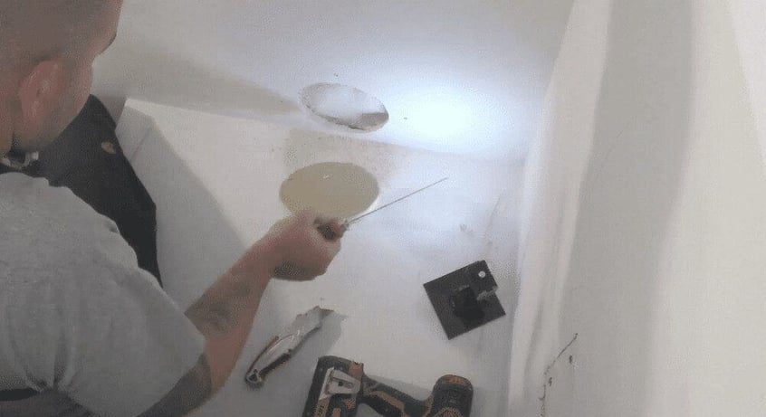 A man cutting a hole out of drywall approximately 3 inches in diameter. His drill and box cutter are on the floor next to him.