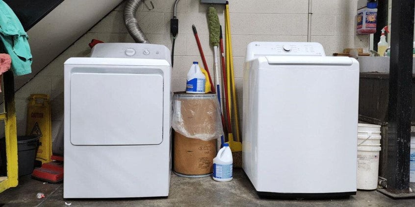 A white washer and dryer installed on the floor of an industrial factory. Various cleaning solutions and implements are stacked around the machines.