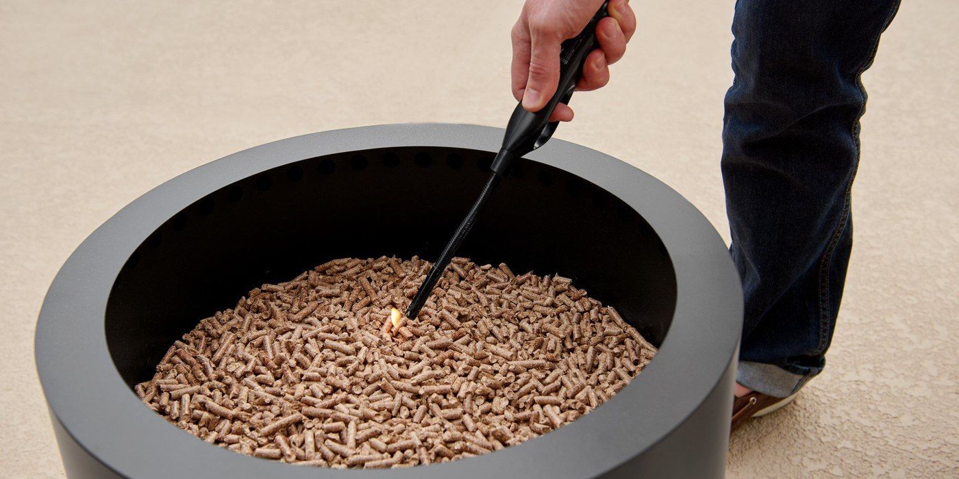 A person reaching into a smokeless fire pit filled with pellets with a long lighter to light a fire