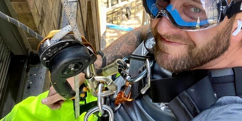 A pest control operator hanging off the side of a building. He is wearing goggles and is strapped into a safety harness. He has a beard and tattoos.