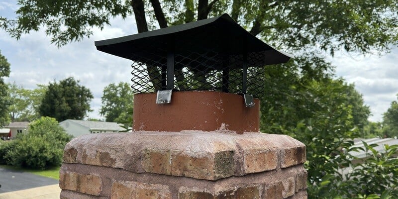 A Shelter adjustable chimney cap installed on a square clay masonry flue. Trees and other houses are visible in the background.