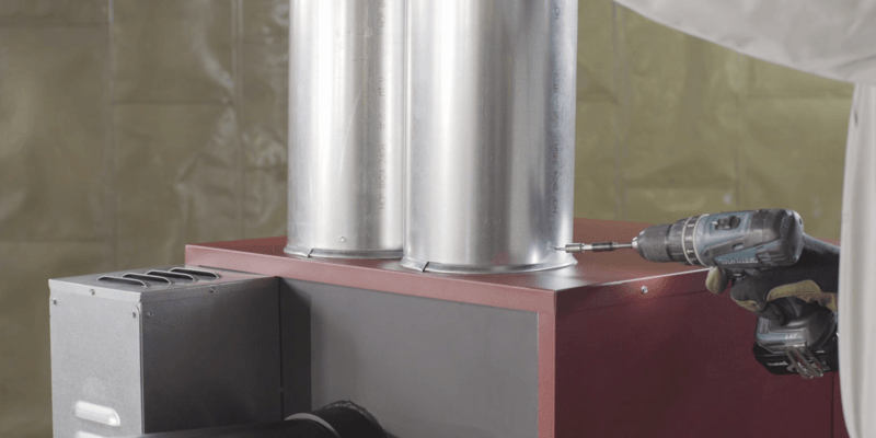 A man installing two duct pipes on the plenum of a Fire Chief FC1000E Wood Burning Furnace.