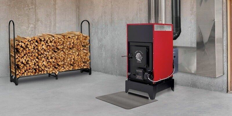 A Fire Chief FC1000E Wood Burning Furnace installed in a basement. Next to the furnace is a log rack filled with wood and a steel stove board.