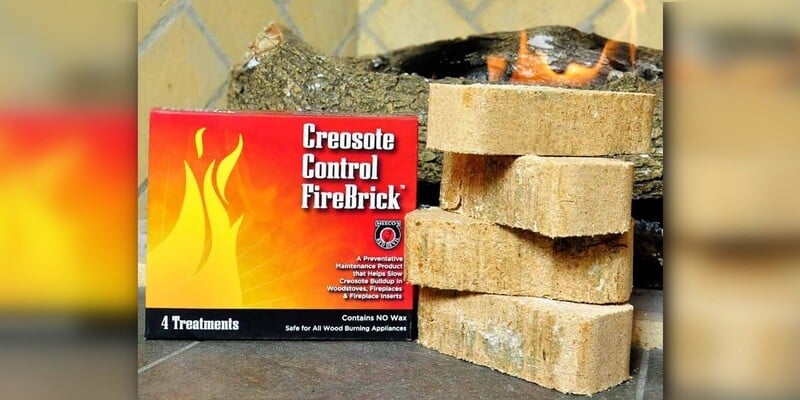 Four Creosote Control FireBricks stacked next to their bright-orange packaging on a hearth. A fire burns in a fireplace in the background.