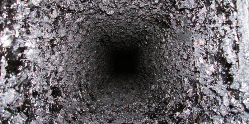 A bird's eye view of a square chimney flue caked in a thick layer of creosote. The bottom of the flue is not visible; instead, there is a black void.