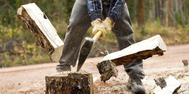 A man splitting a log in two with an ax. He is wearing yellow gloves, gray pants, and a blue flannel.