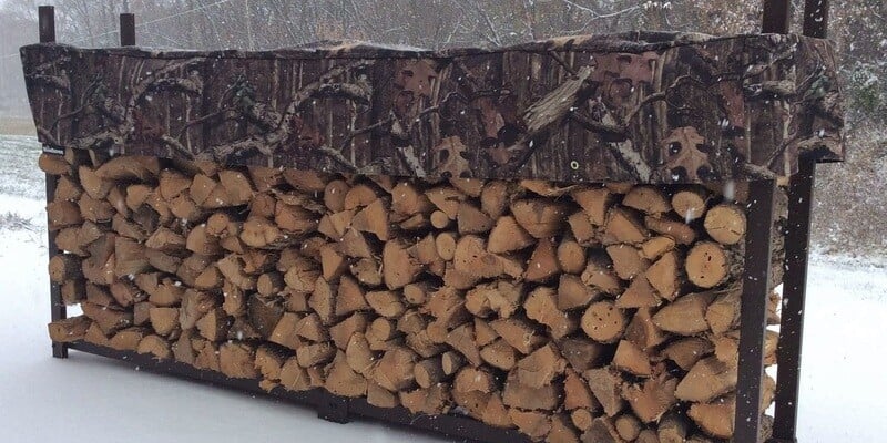 A rack full of firewood out in the snow. The rack is covered with a camouflage cover. 