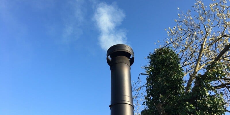 A ground-up shot of a black stove pipe emitting white smoke. Trees can be seen to the right of the pipe, all against a bright blue sky.