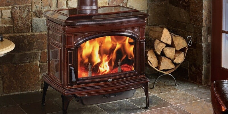 A wood stove resting on a tile floor with a stone wall behind it. A bundle of firewood rests next to the stove.