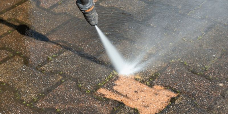 A pressure washer water nozzle spraying water to clean a brick patio. One of the breaks has been deeply cleaned by the pressure washer.