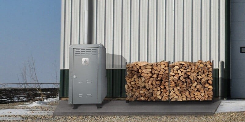 An Ignite Tech HD90X Outdoor Wood Furnace installed on a concrete slab outside a business. It sits next to a firewood rack full of wood.