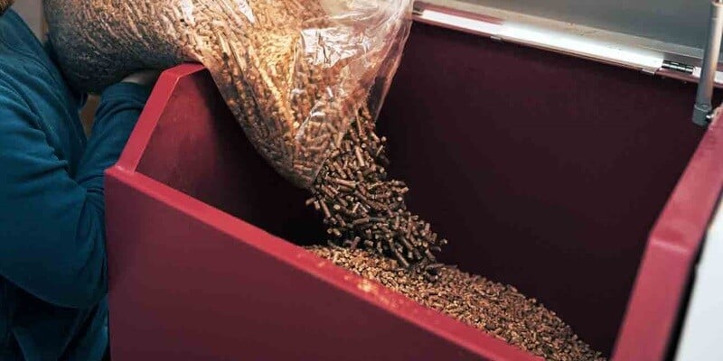 A man pouring a large, clear bag of pellet fuel into the hopper of a wood pellet stove.