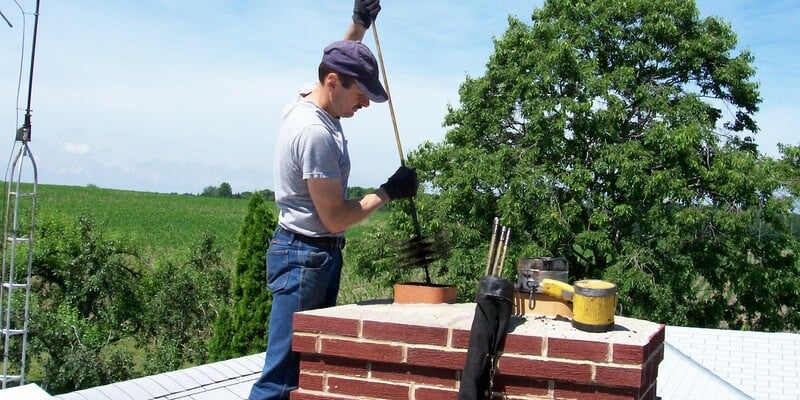 A chimney sweep atop a roof. He is inserting a chimney sweep's brush in one of the flues on a multi-flue chimney.