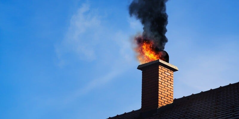 A masonry chimney with a chimney fire roaring out of the flue. The roof and a bright blue sky are visible in the background.