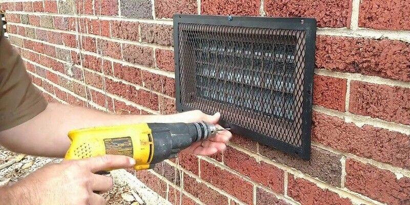 A man installing a HY-GUARD EXCLUSION foundation vent cover over a foundation vent on a home with brick siding.