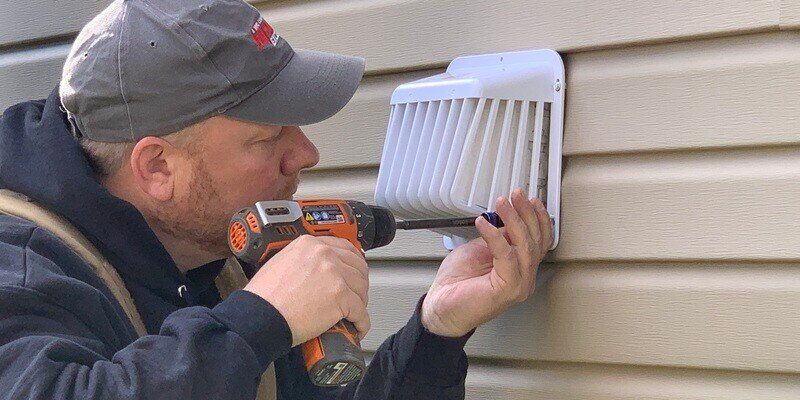 A man using an orange drill to affix a HY-GUARD EXCLUSION plastic dryer vent cover to the side of a house with beige siding.