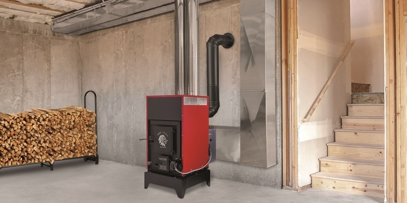 A Fire Chief FC1000E wood burning furnace sitting in an unfinished basement. A firewood rack full of wood rests to the furnace's right.