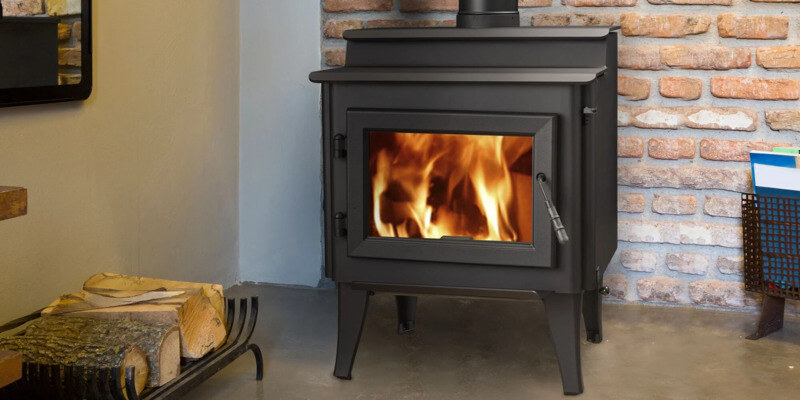 A wood stove installed in a home. Its back rests against a brick wall. A rack with a few firewood logs sits to the stove's right.