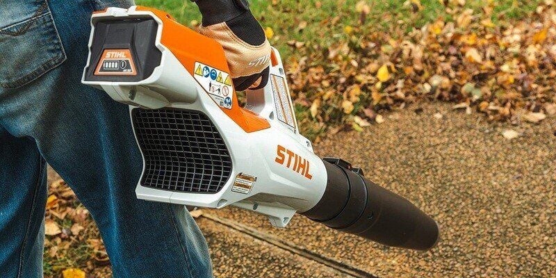 A person in blue jeans using a STIHL BGA 57 leaf blower to blow leaves off of a sidewalk.