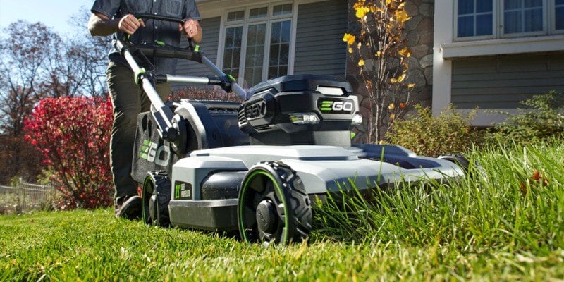 A man using an EGO electric lawn mower to mow his lawn. The mower is actively rolling over some tall grass.