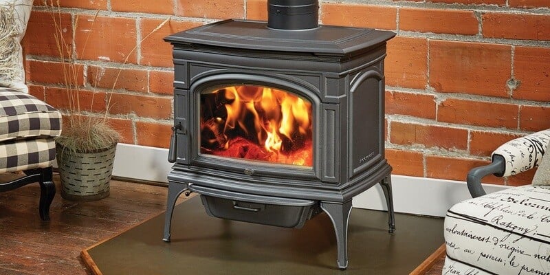 A Lopi Stoves Rockport NexGen-Hybrid wood stove installed on a hearth pad in a living room between two chairs. The wall behind it is made of brick.