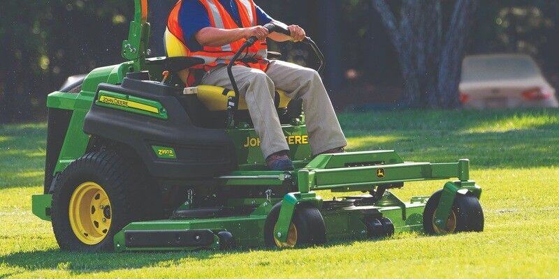 A man in an orange high-visibility vest riding a John Deere zero-turn mower, cutting grass in a field.