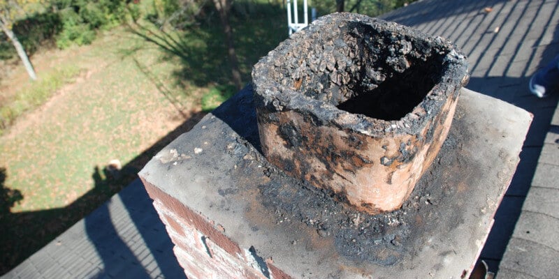 An uncapped masonry chimney flue covered in creosote. A sloped roof and the home's yard are visible in the background.