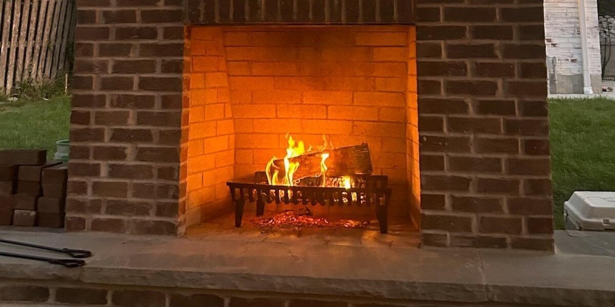 An outdoor fireplace with a fire burning in the firebox. The logs are burning on top of a Franklin G Series fireplace grate.