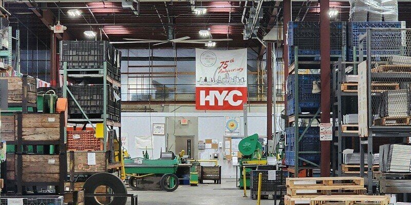 A wide-angle shot of the HY-C factory floor with shelves stacked high with raw metal materials. A large HY-C banner hangs on the far wall.