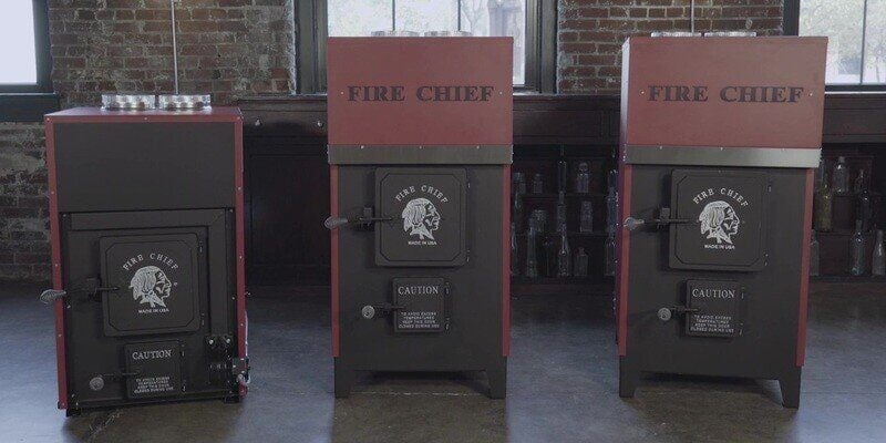 Three Fire Chief wood burning furnace models sitting on a concrete floor. A brick wall with windows can be seen in the background.