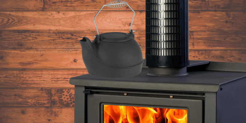 A wood stove kettle steamer resting on top of a wood stove with a fire burning in its firebox. The wall in the background features wood paneling.